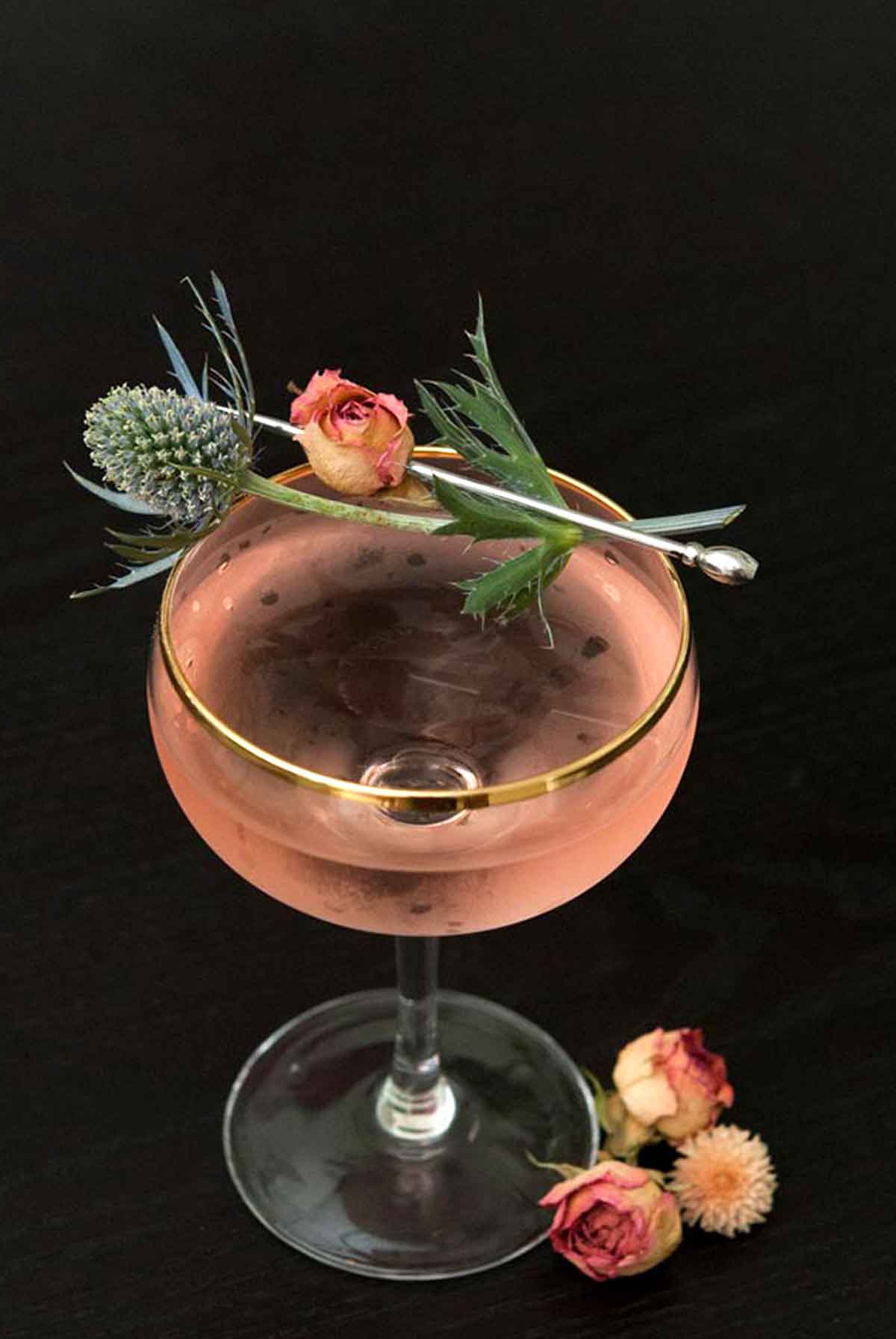 A pink martini, garnished with a pink spray rose and a sprig of thistle on a black table.