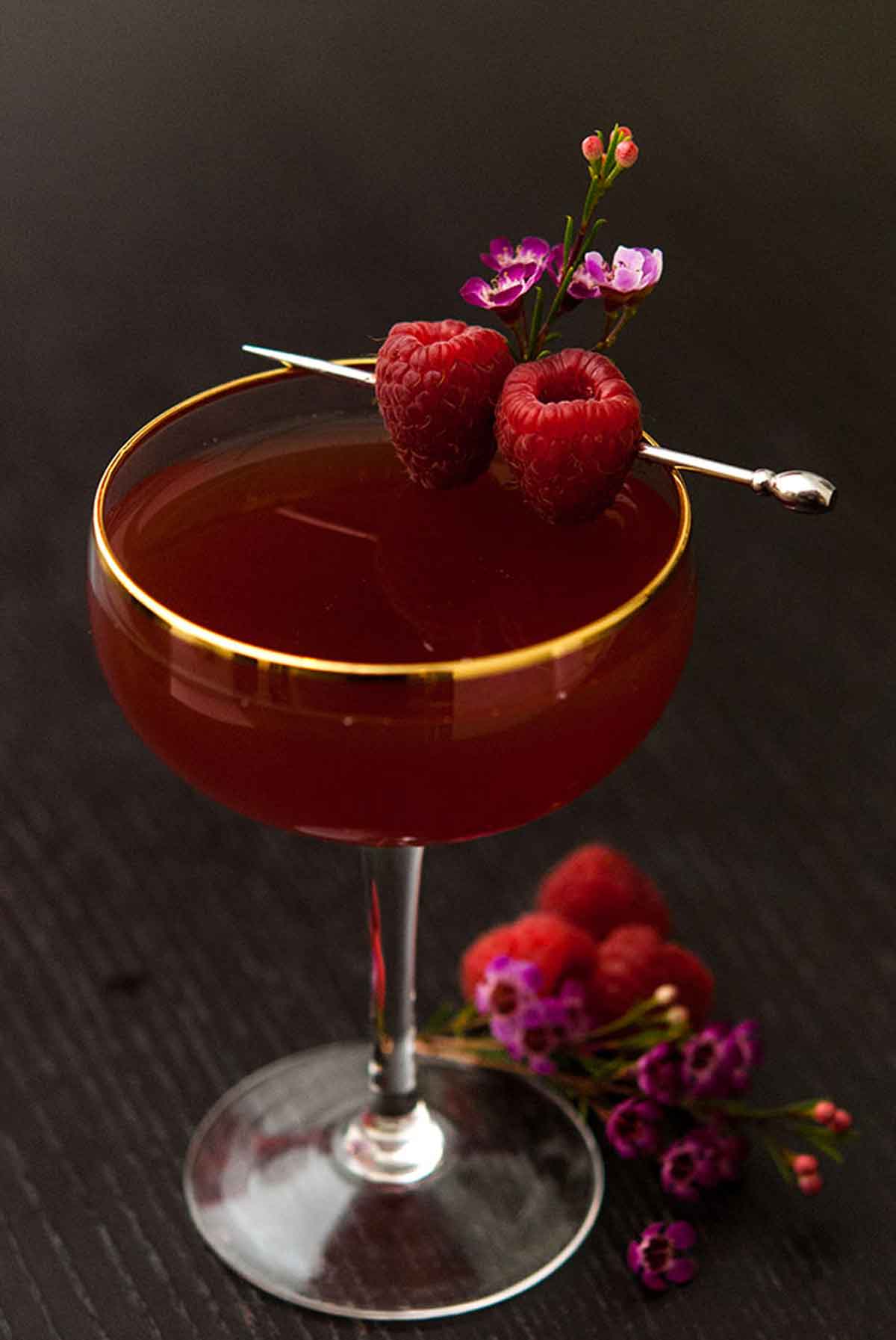 A cocktail on a dark table garnished with raspberries and small flowers, with a few raspberries and flowers at its base.