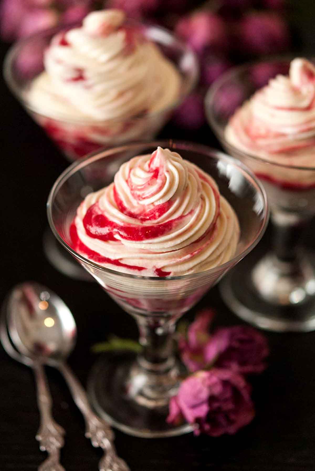 3 glasses of white chocolate mousse with raspberry compote swirls on a black table with a few roses and spoons at their base.