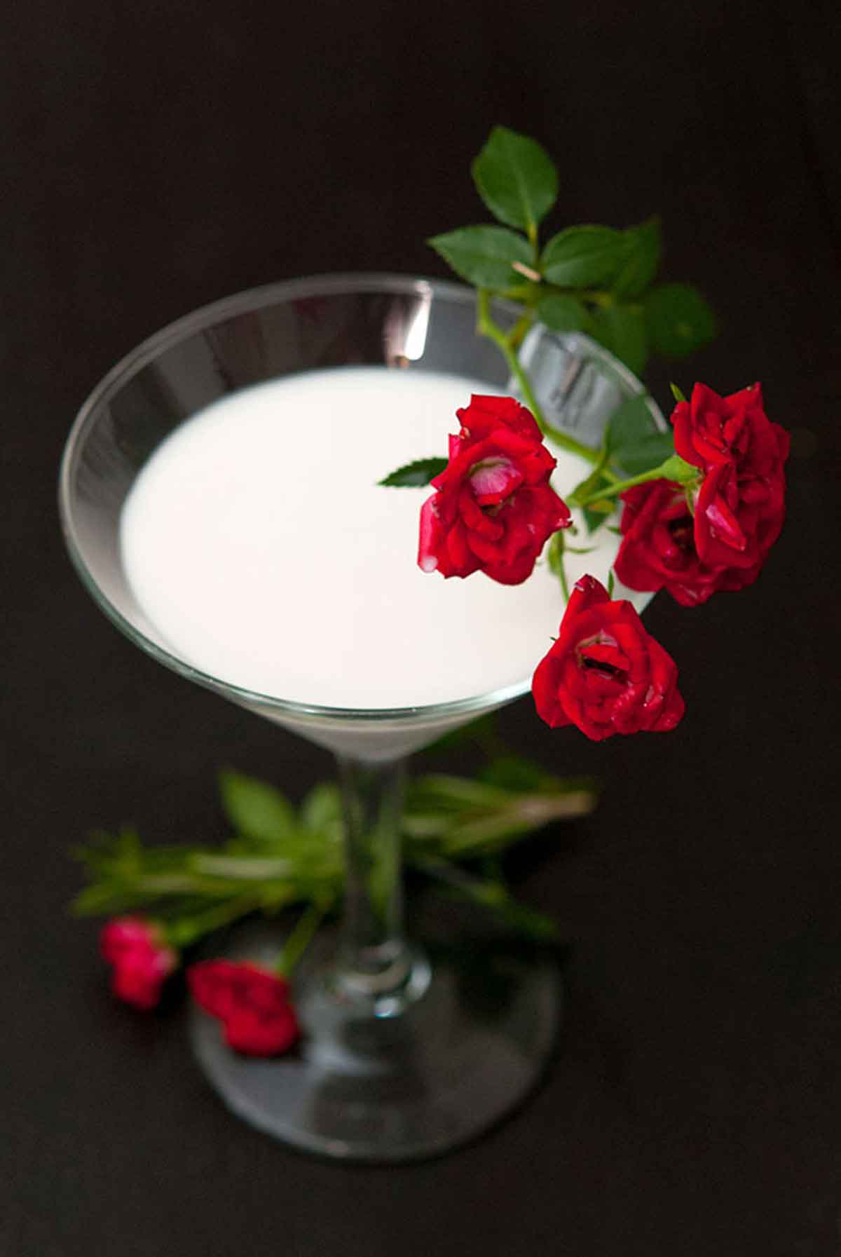 A cocktail in a martini glass on a black table, garnished with a few small spray roses.