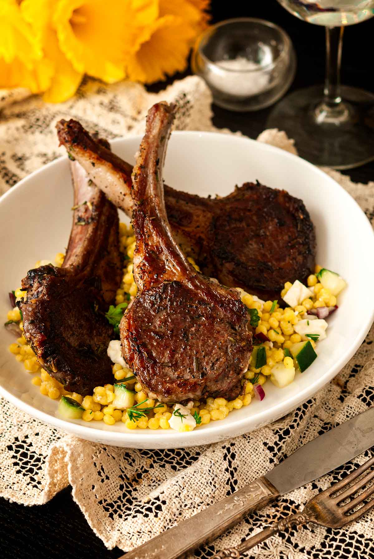 3 seared lamb chops in a bowl with pearl couscous on a table cloth, with flowers in the background.