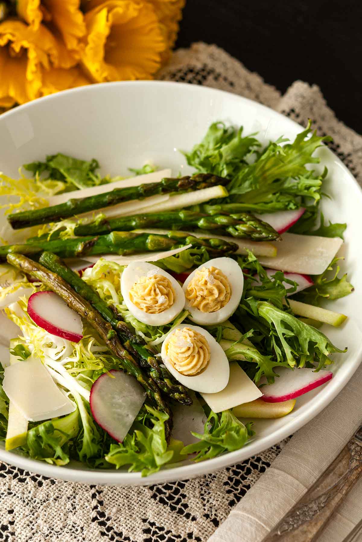 A salad in a bowl on a lace table cloth, topped with small deviled eggs with flowers in the background.
