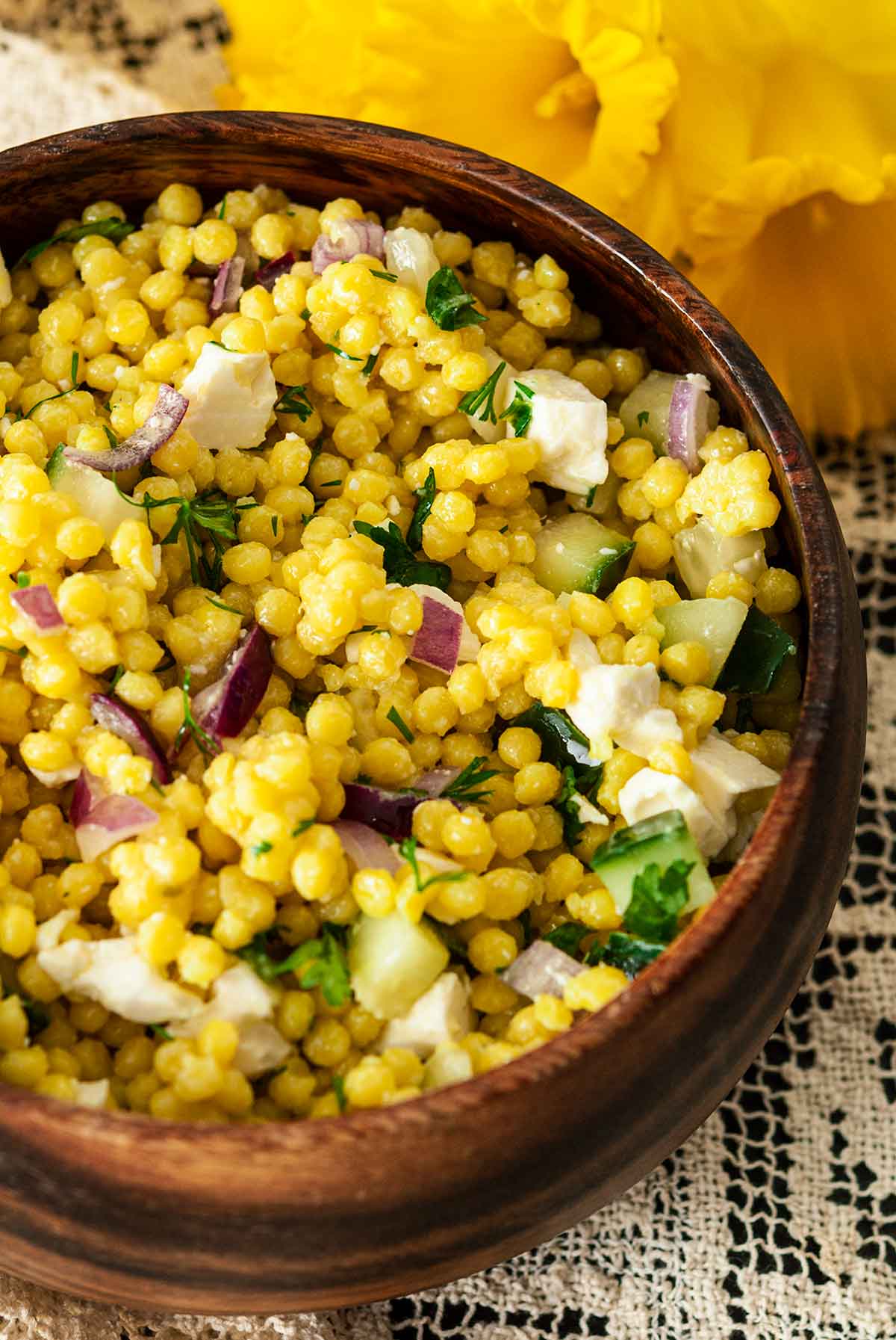 A wooden bowl of turmeric pearl couscous beside flowers on a lace tablecloth.