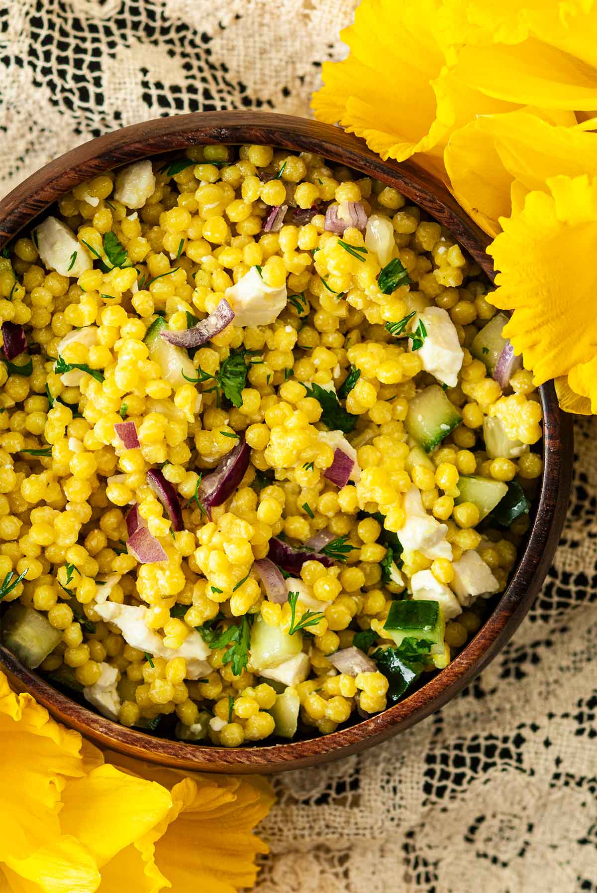 A wooden bowl of turmeric pearl couscous beside flowers on a lace tablecloth.