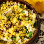 A wooden bowl of turmeric pearl couscous beside flowers on a lace tablecloth.