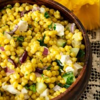 A wooden bowl of turmeric pearl couscous beside flowers on a lace tablecloth.