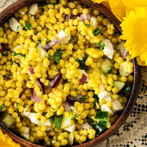 A wooden bowl of turmeric pearl couscous beside flowers on a lace tablecloth.