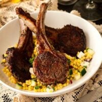 3 seared lamb chops in a bowl with pearl couscous on a table cloth, with flowers in the background.