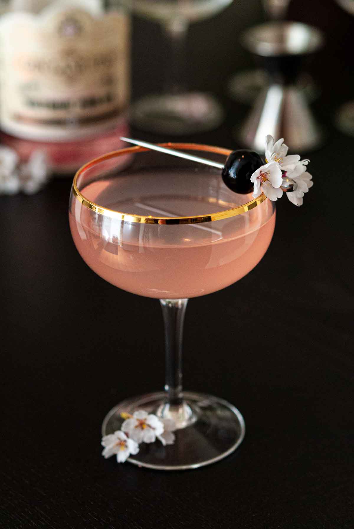 A pink cocktail in a coup glass, garnished with a cherry and flowers, on a table.