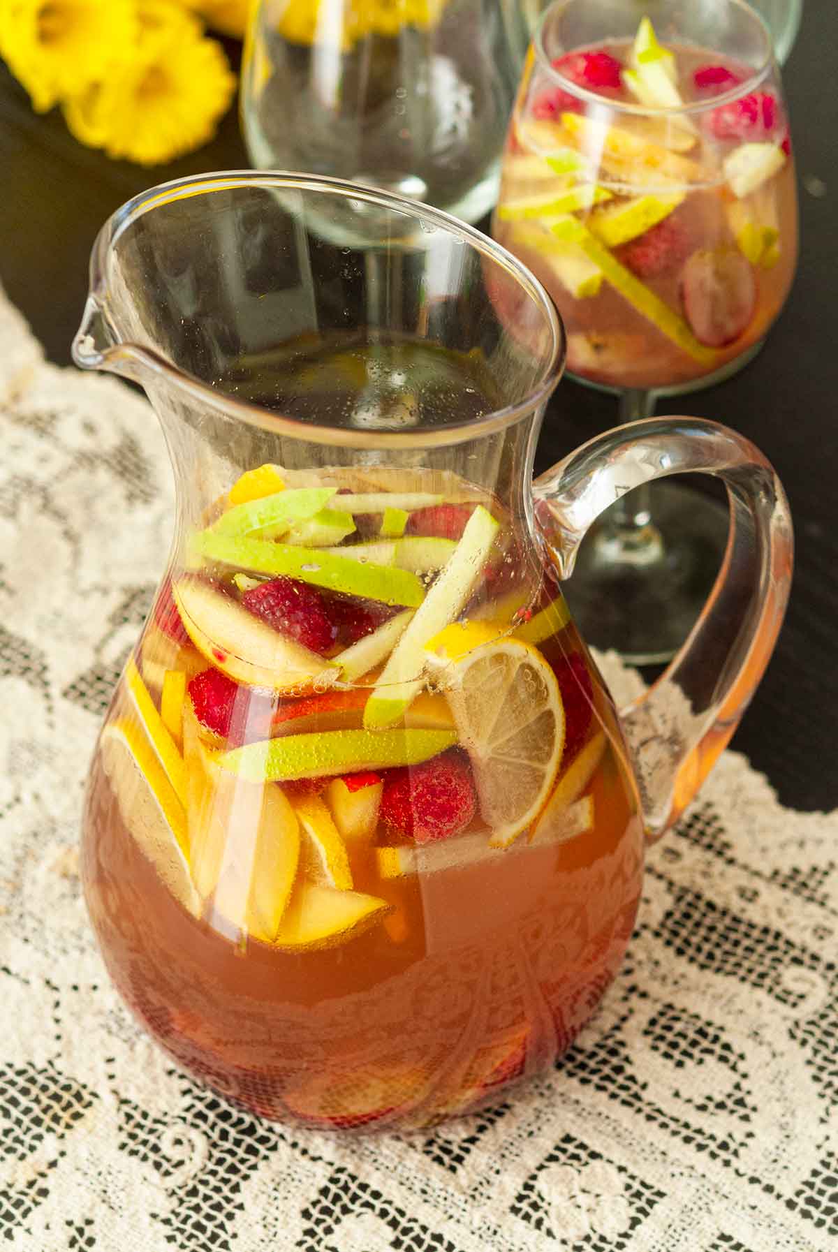 A pitcher of sake sangria on a table cloth, beside glasses and flowers.