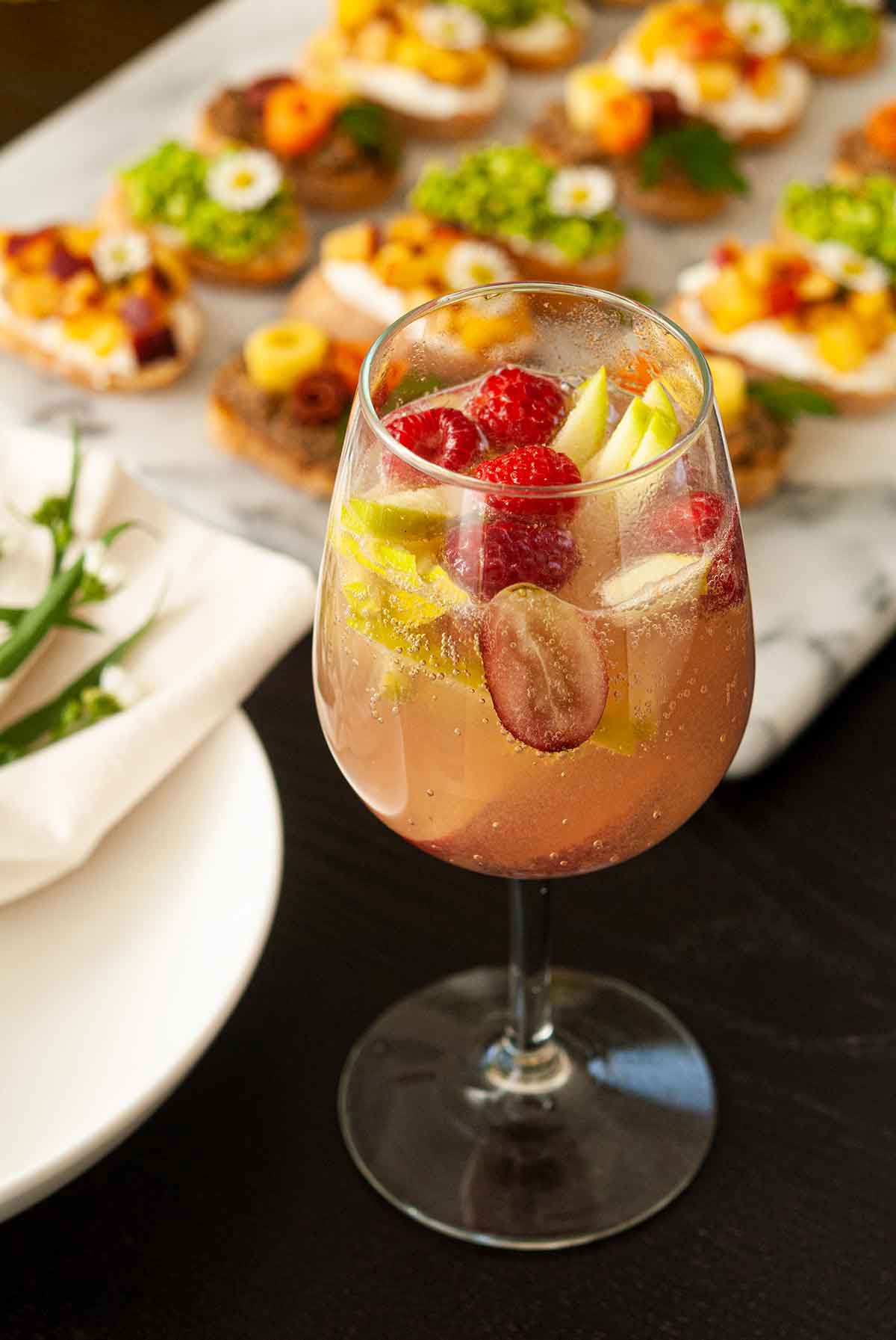 A glass of sake sangria, full of fruit, on a table beside appetizers.