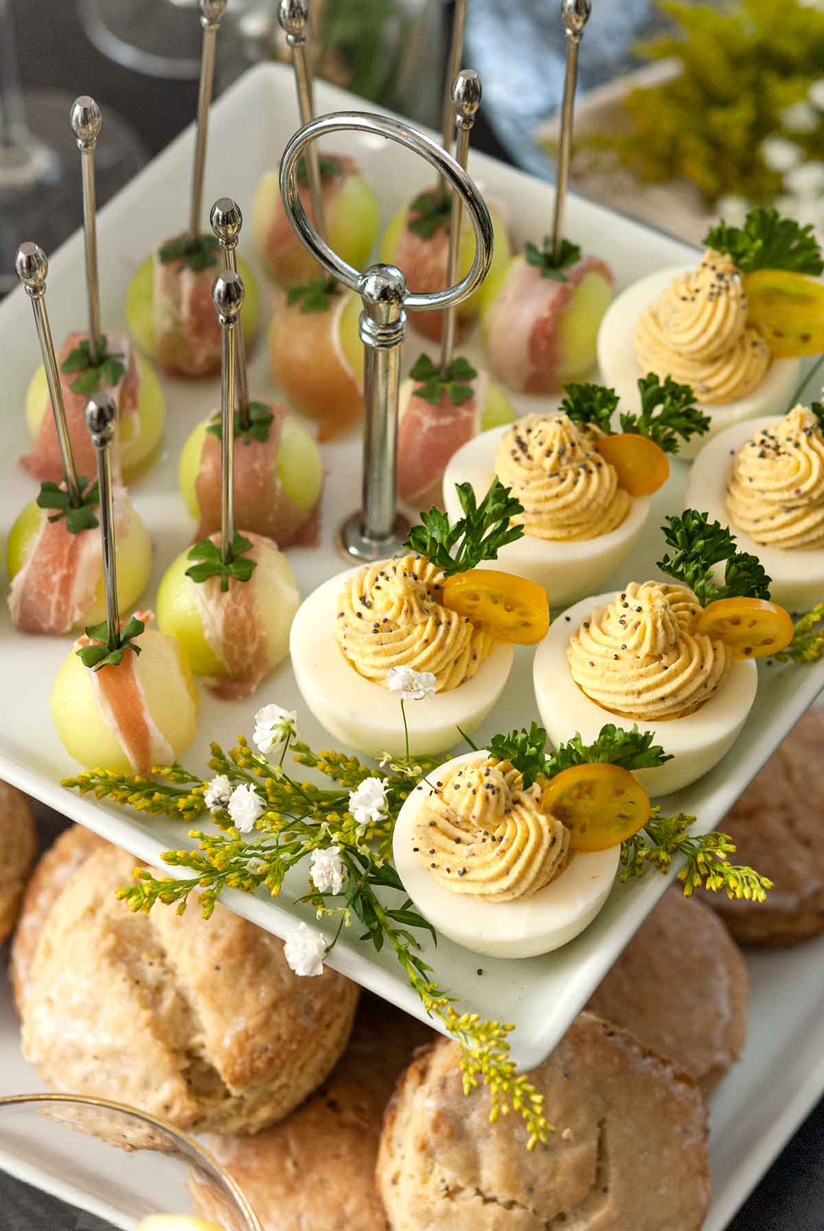 An appetizer tier with deviled eggs and prosciutto-wrapped melon above scones on a table.