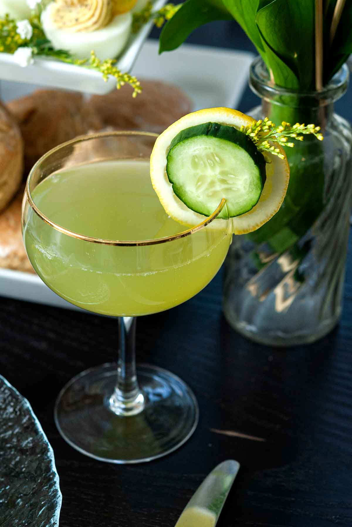 A cocktail garnished with cucumber and lemon on a table in front of a tier of sones and deviled eggs.