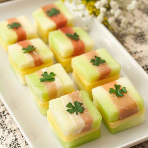 8 St. Patrick's Day melon appetizers with clovers on top, on a white plate, on a lace table cloth..