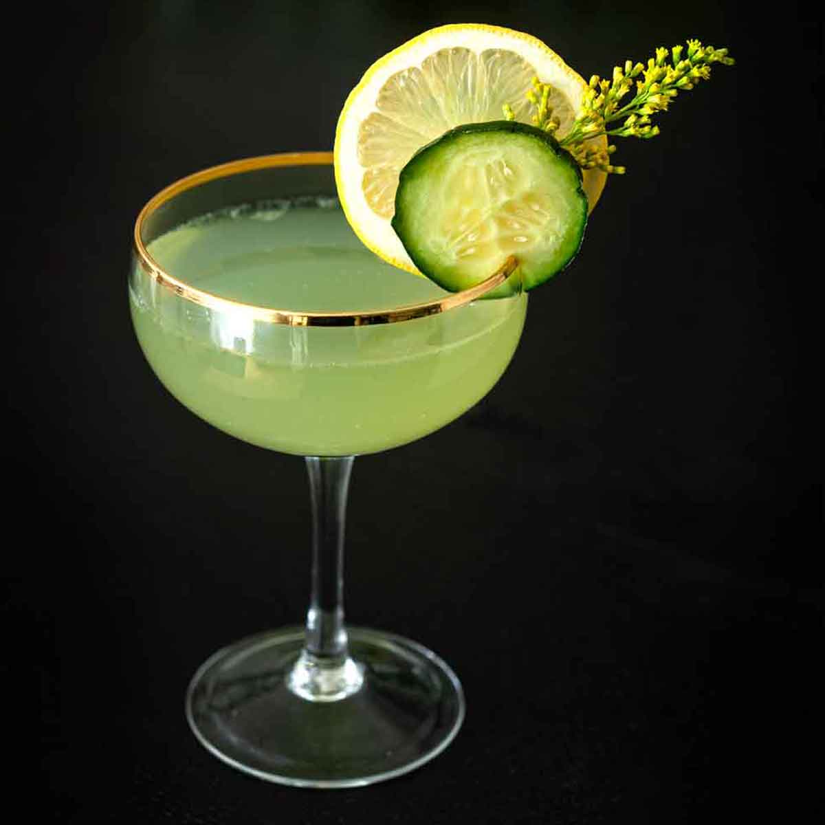 A cocktail in a coup glass, garnished with a cucumber slice, lemon, and wispy flower on a table.