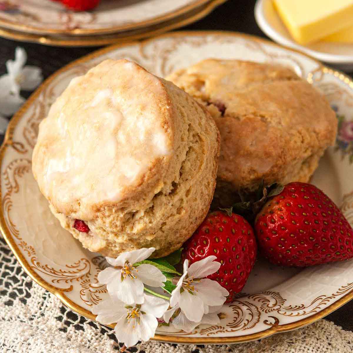 2 scones on a decorative plate with flowers and strawberries, in front of stacked plates and more strawberries.