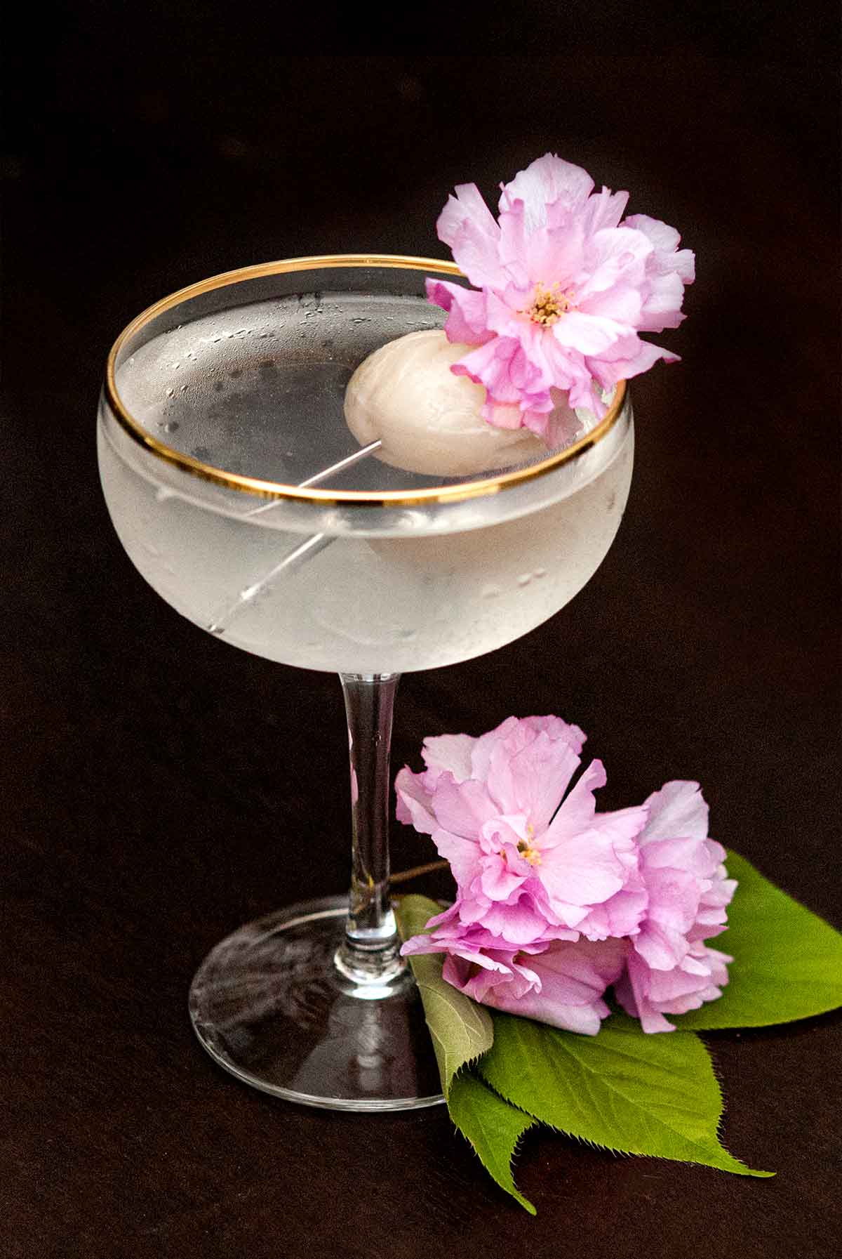 A cocktail garnished with a lychee and cherry blossom flowers, with flowers and a few leaves at its base.