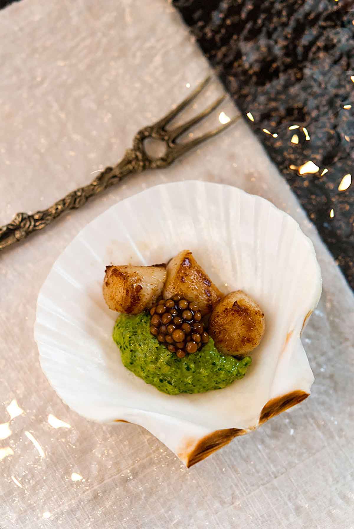 A small shell on a plate containing 3 small scallops, pesto cream and mustard caviar.