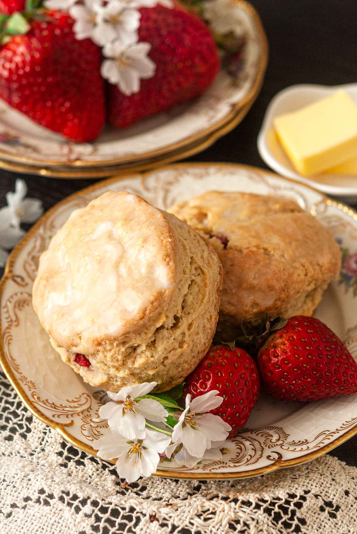 2 scones on a decorative plate with flowers and strawberries, in front of stacked plates and more strawberries.