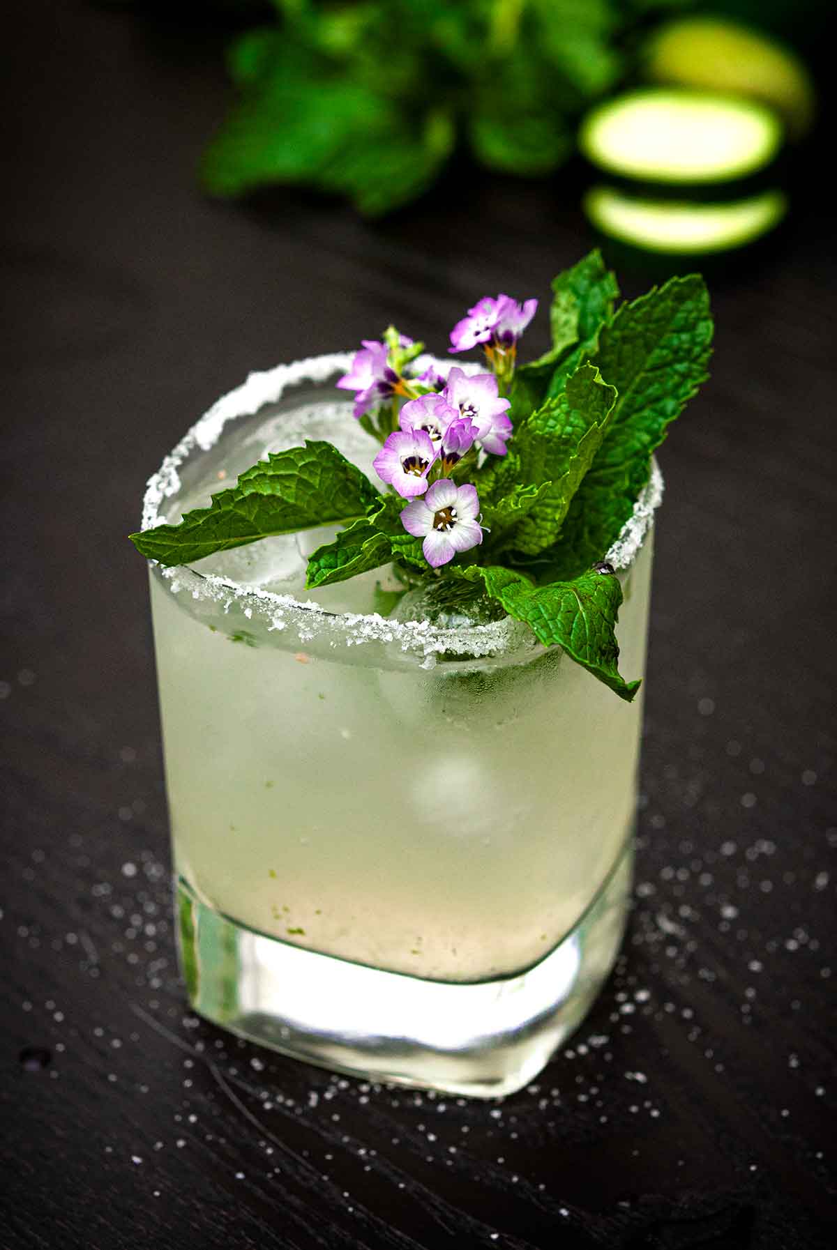 A cucumber mint margarita, garnished with small flowers and mint, on a table lightly sprinkled with salt.