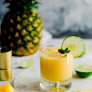 A pineapple margarita surrounded by lime and pineapple slices, in front of a pinapple.