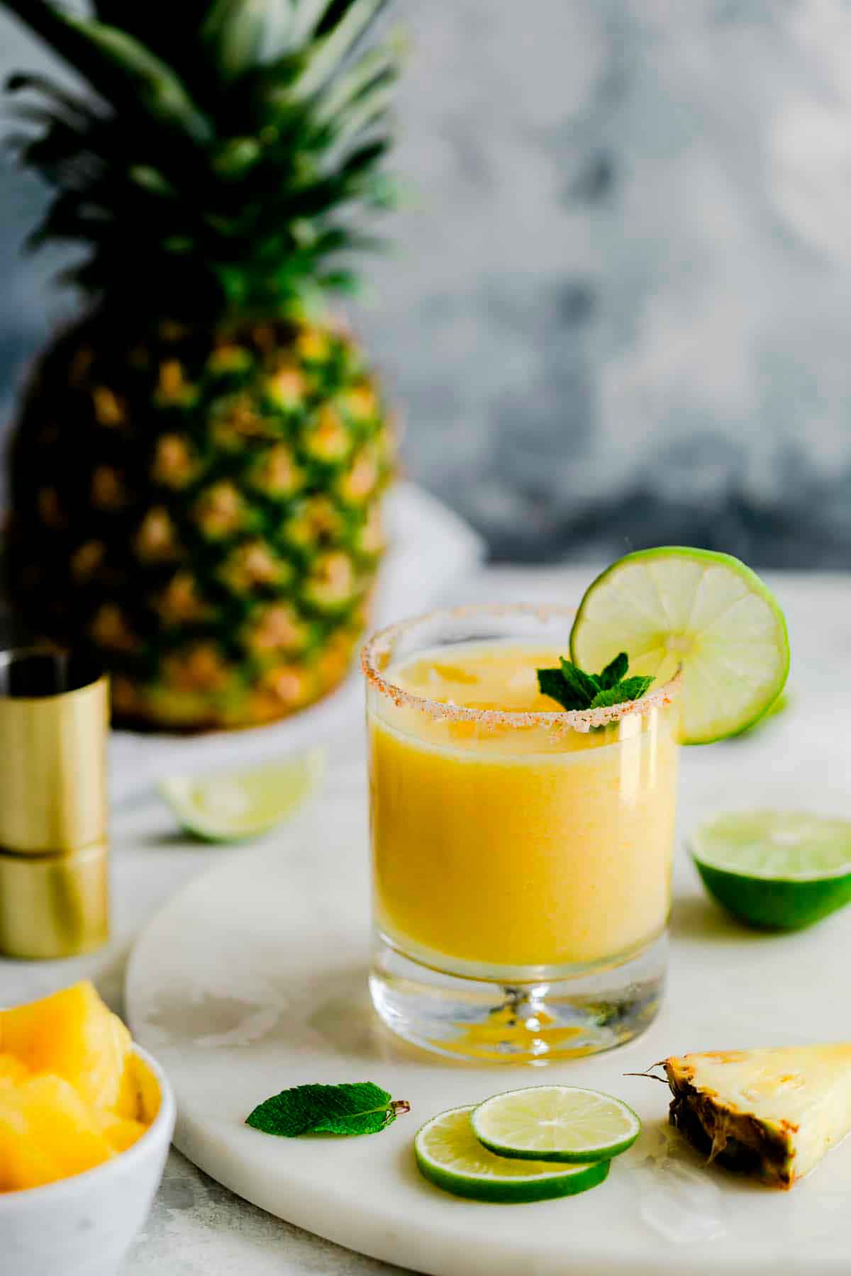 A pineapple margarita surrounded by lime and pineapple slices, in front of a pinapple.