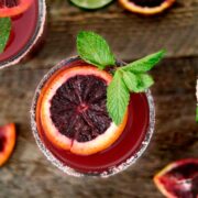 A blood orange margarita, garnished with blood orange and mint, on a table with citrus slices.