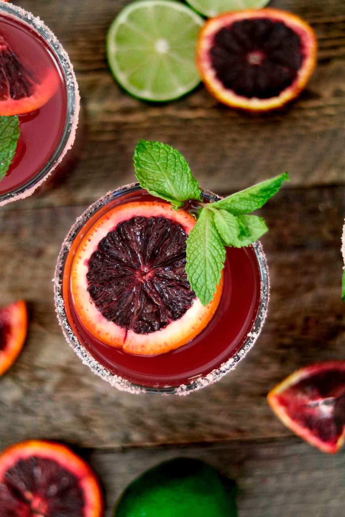 A blood orange margarita, garnished with blood orange and mint, on a table with citrus slices.