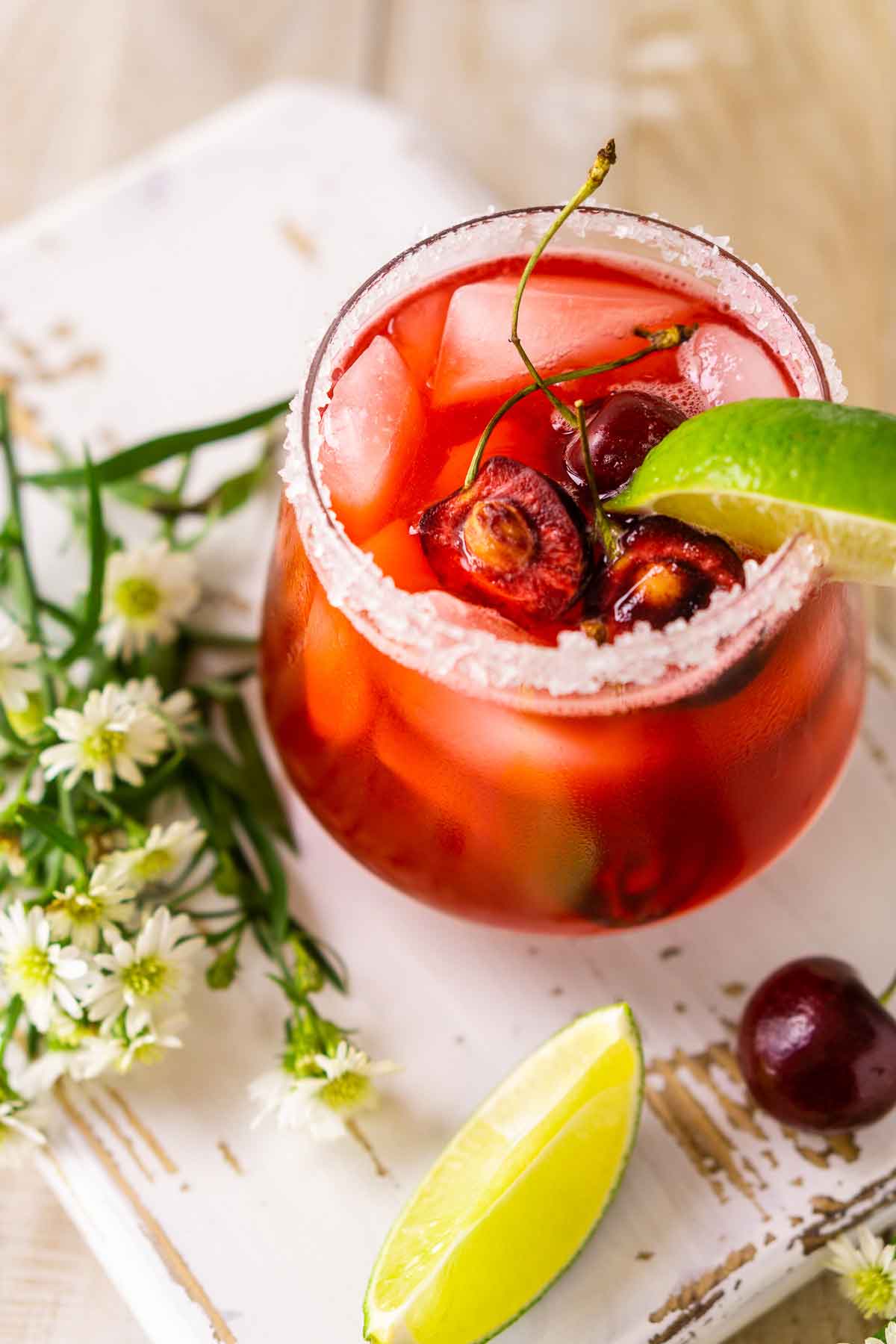 A cherry margarita, rimmed with salt, on a table beside flowers and lime slices.