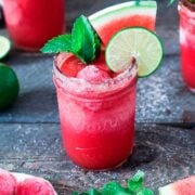 A watermelon margarita on a table, garnished with watermelon, lime and mint, surrounded by others.
