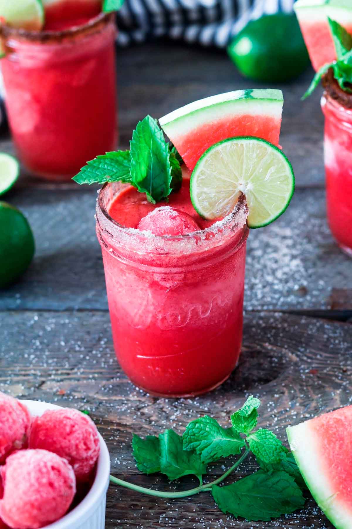 A watermelon margarita on a table, garnished with watermelon, lime and mint, surrounded by others.