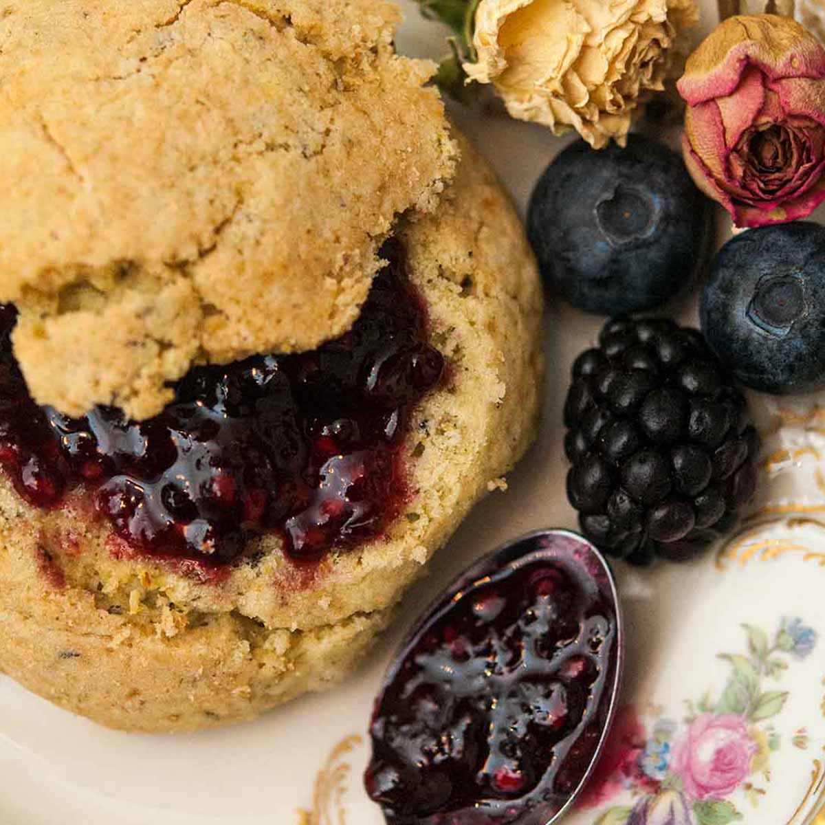 A scone with jam on a decorative plate next to a spoon with jam, a few berries and a few dry roses.