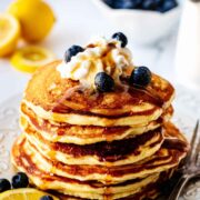 A stack of pancakes topped with cream and blueberries, with slices in the backgroun.
