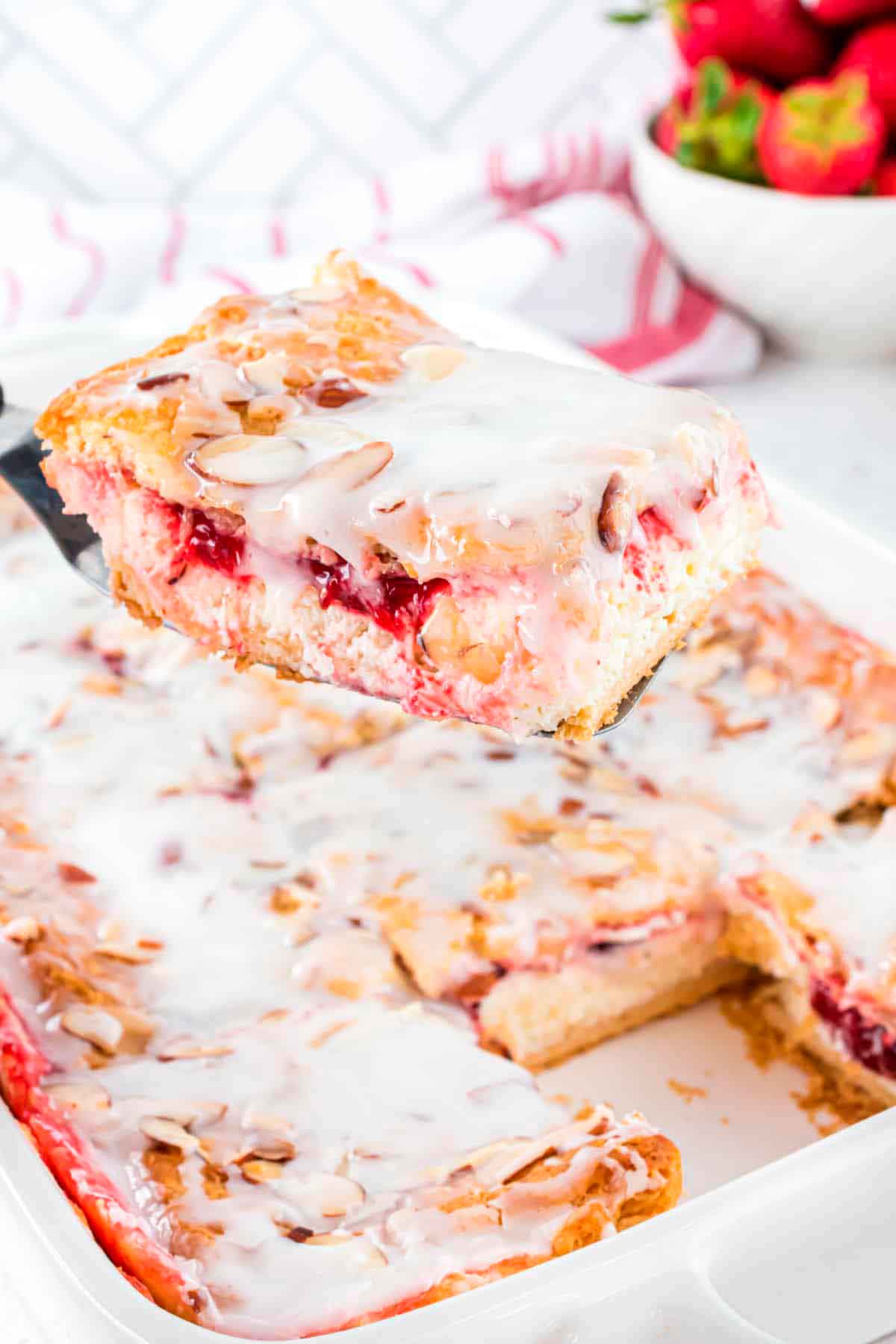 A spatula holding up a strawberry cheesecake danish above a full danish in a baking dish.