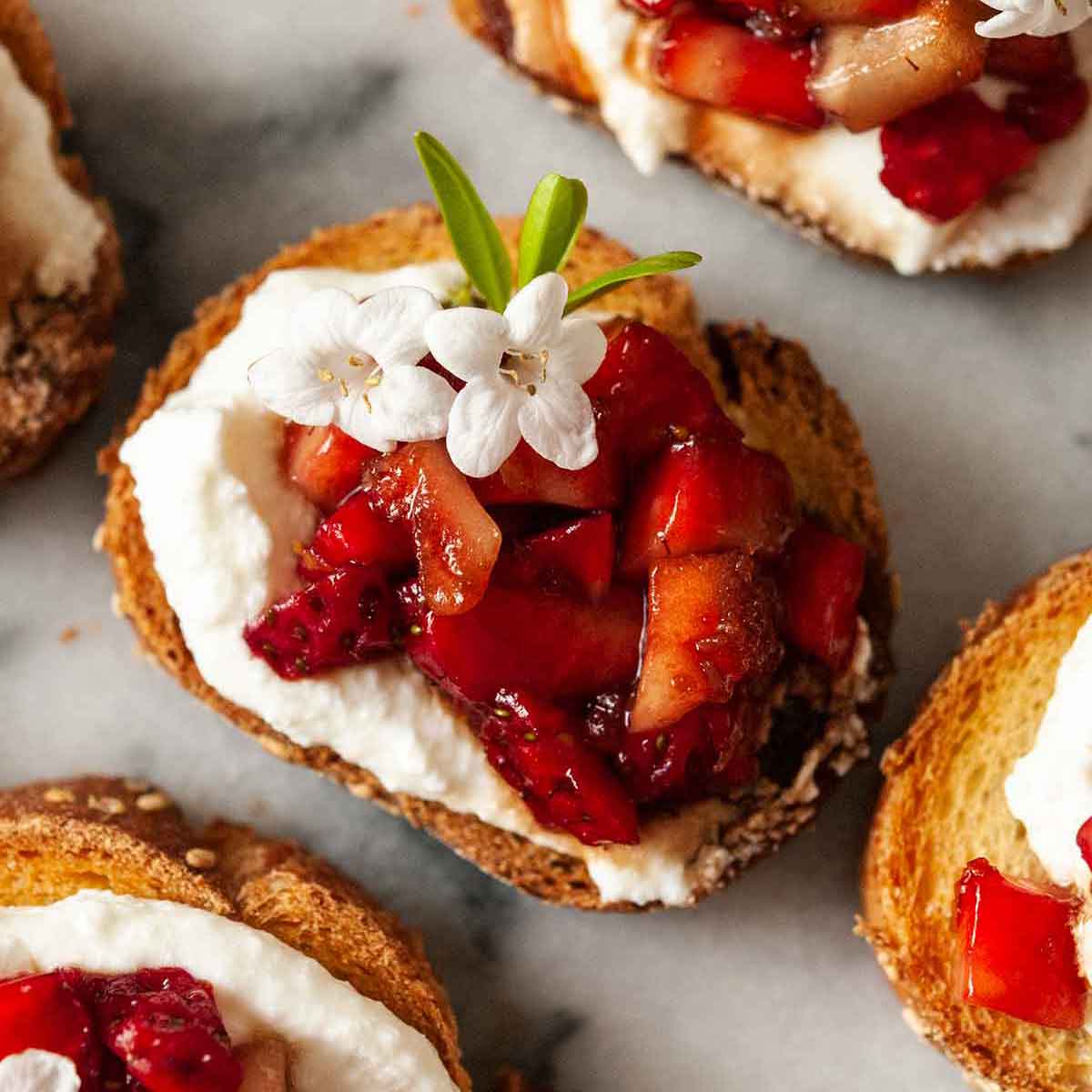 A balsamic strawberry crostini, garnished with a few small flowers, surrounded by others on a marble plate.