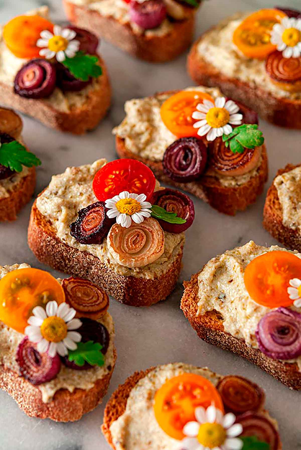 11 crostini garnished with daisies, onions and tomatoes on a marble plate.