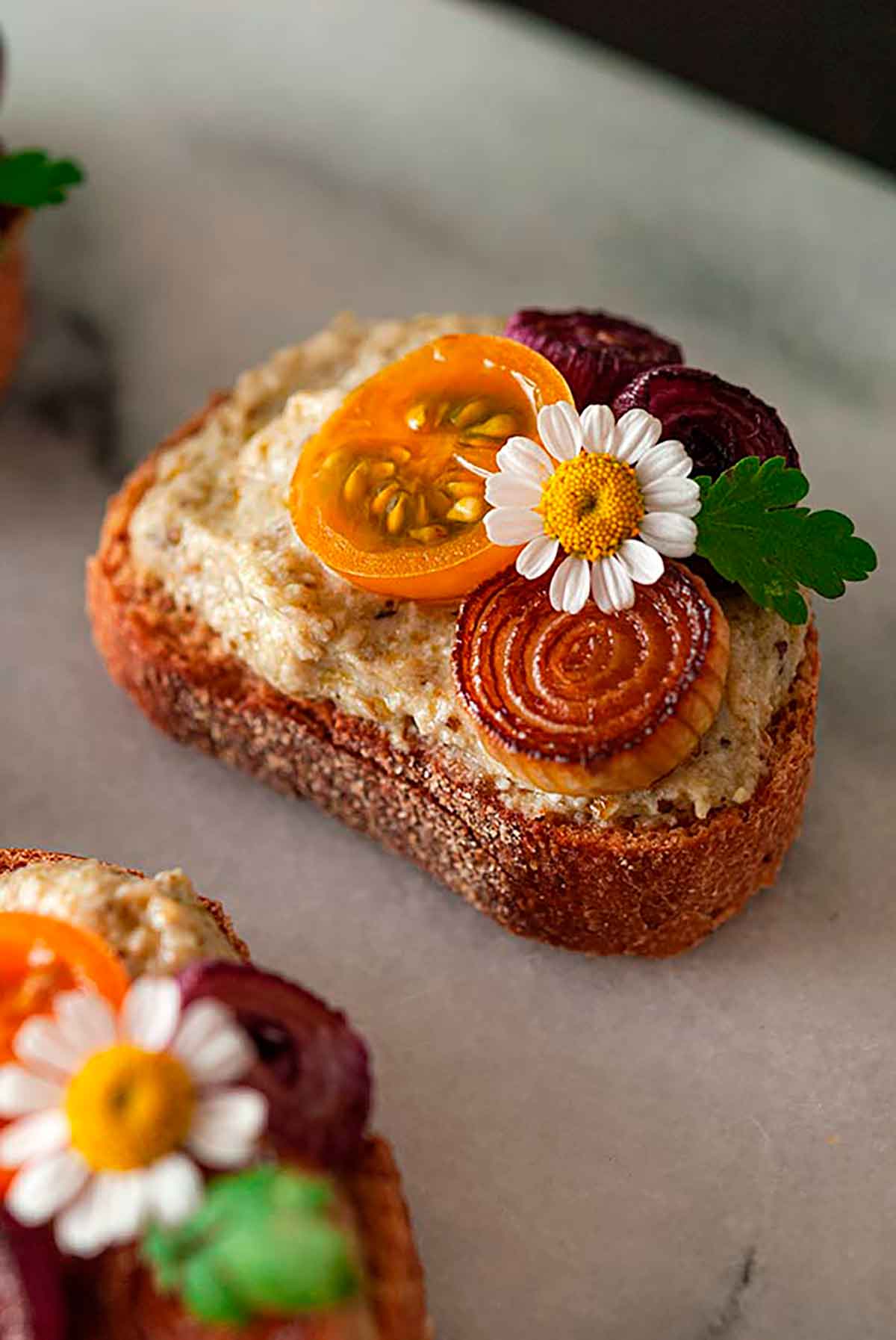 A pesto crostini with onions, tomato and small flower on a marble plate.