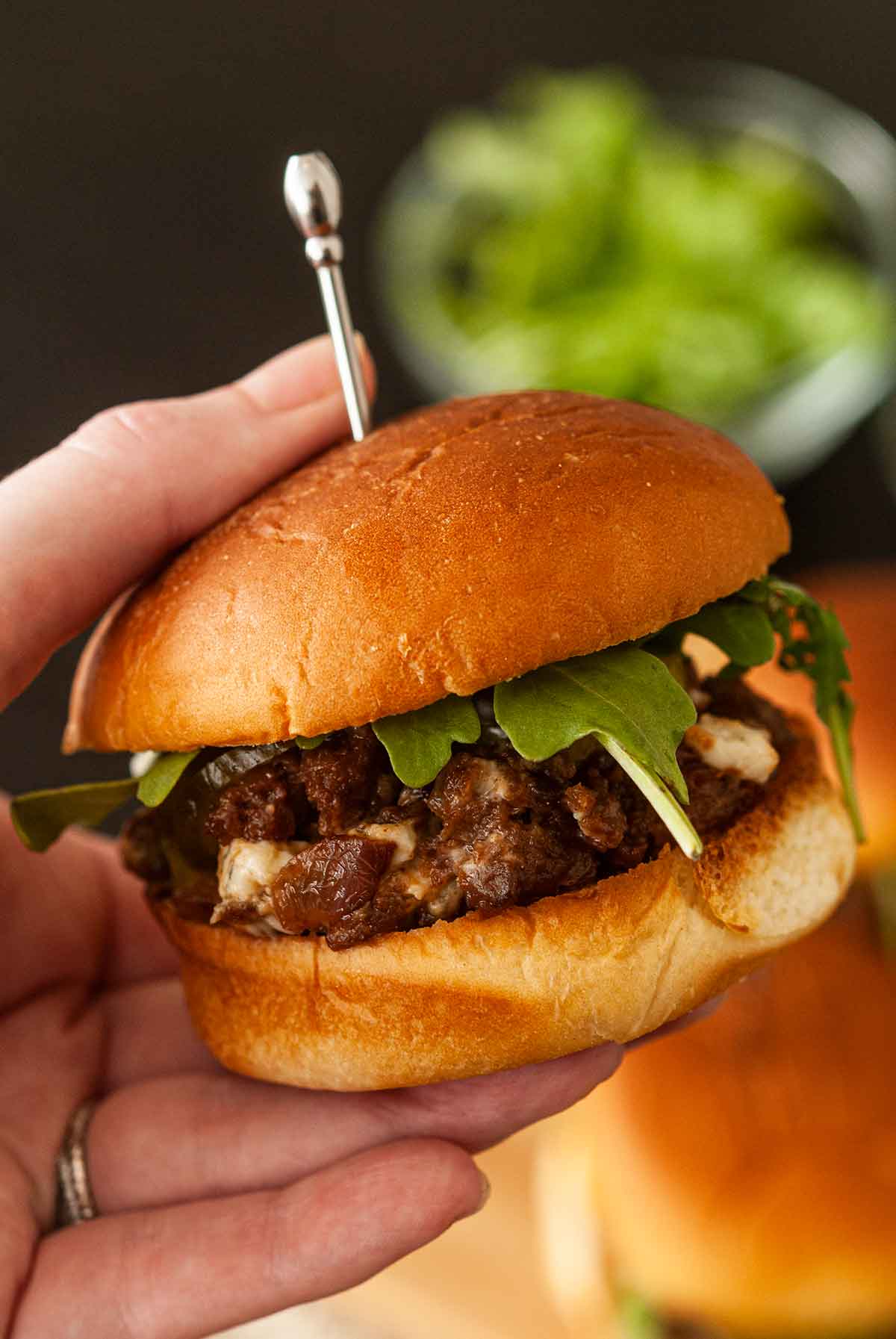 A hand holding a barbecue slider above a table with other sliders and a bowl of greens.