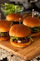 4 barbecue sliders on a cutting board above a lace table cloth with garnishes in the background.