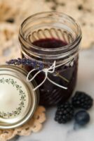 A jar of jam tied with a string with a sprig of lavender tucked into the string with a lace table cloth in the background.