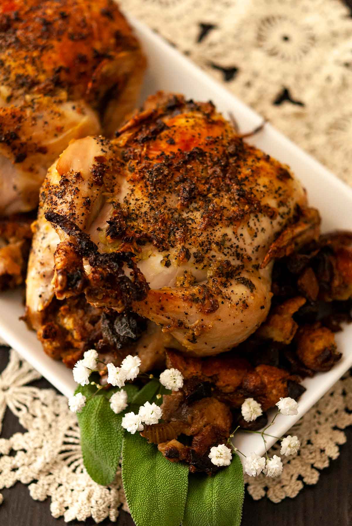 A cooked Cornish game hen on a plate, on top of a lace doily, beside stuffing, a few flowers and 2 sage leaves.