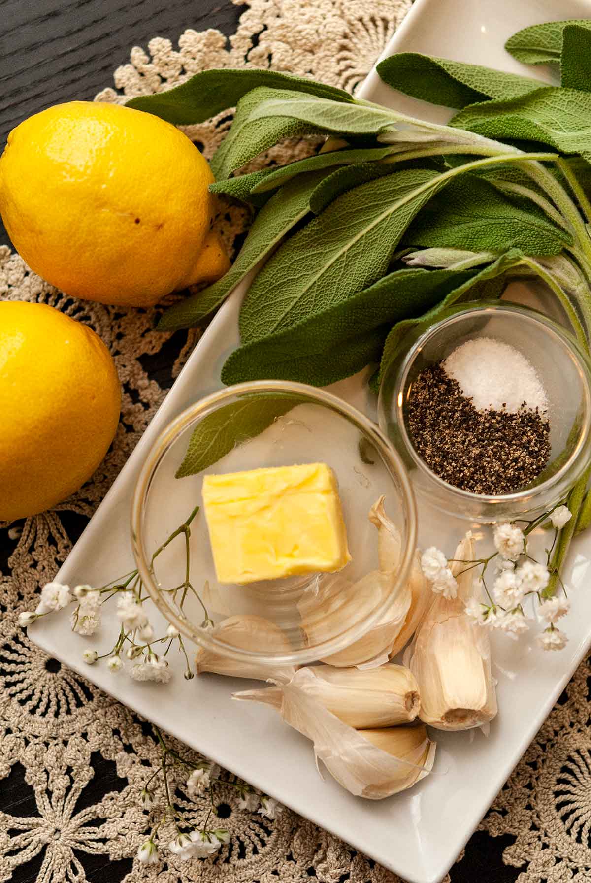 butter, garlic, spices, sage and lemons on a table cloth with a few scattered flowers.