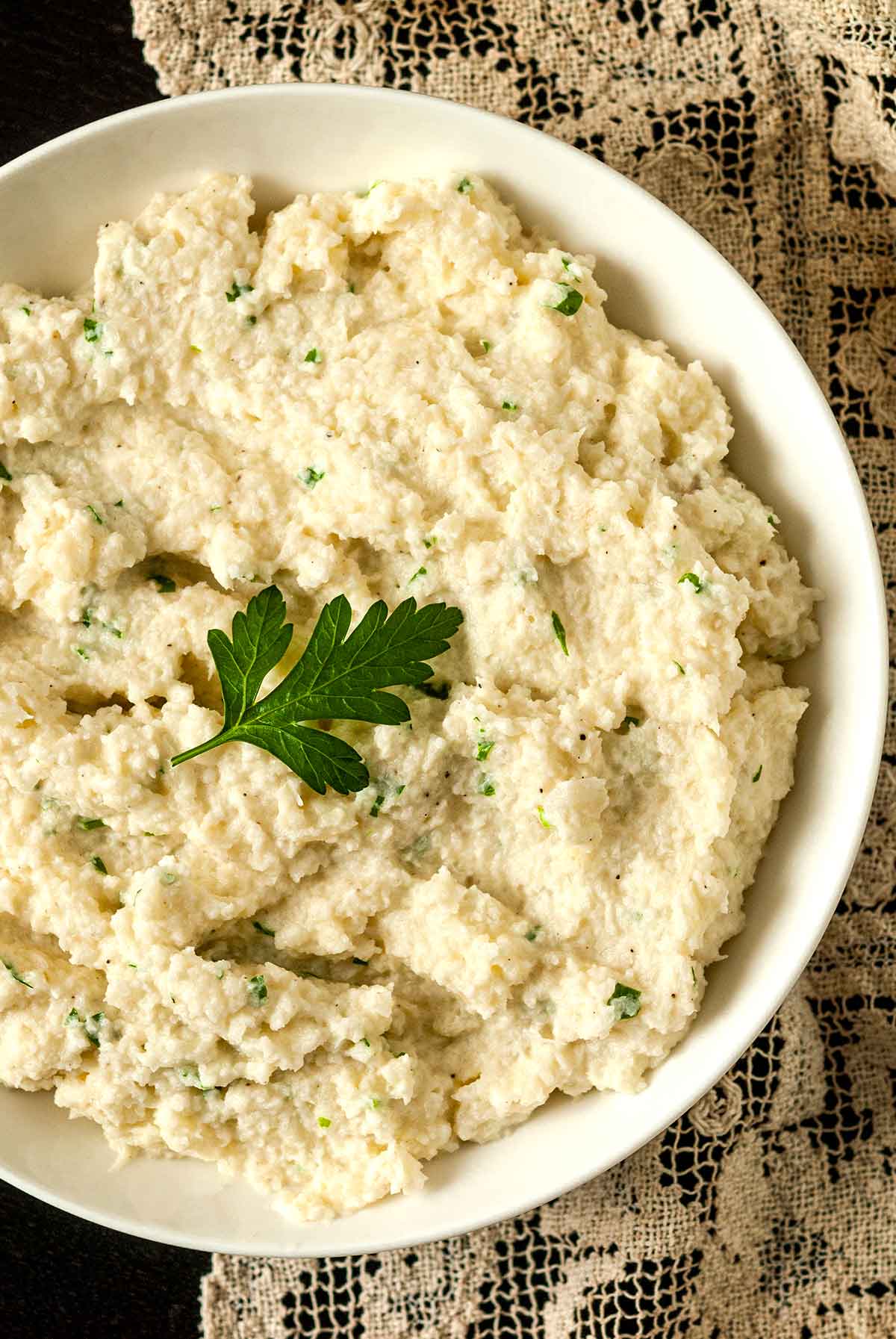 A bowl of garlic-parmesan mashed cauliflower, garnished with a parsley leaf on a lace table cloth.