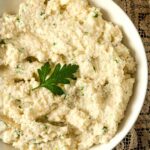 A bowl of garlic-parmesan mashed cauliflower, garnished with a parsley leaf on a lace table cloth.