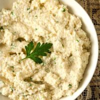 A bowl of garlic-parmesan mashed cauliflower, garnished with a parsley leaf on a lace table cloth.