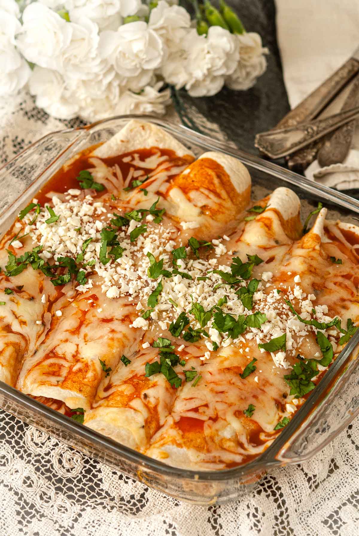 A Pyrex dish with 4 chicken enchiladas on a lace tablecloth with flowers and silverware in the background.