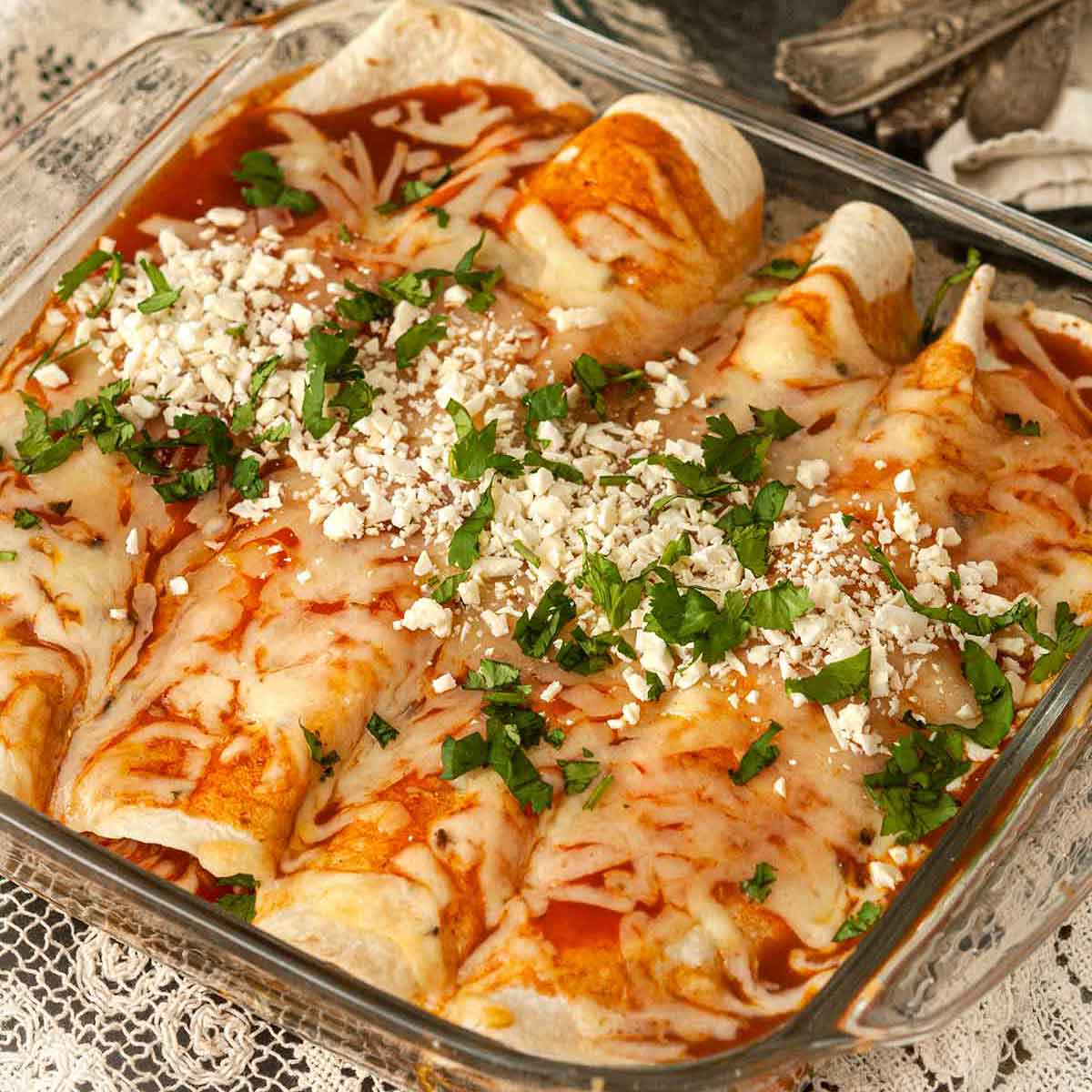 A Pyrex dish with 4 chicken enchiladas on a lace tablecloth with flowers and silverware in the background.