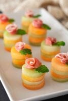 8 small melon appetizers with a prosciutto rose and mint leaves on top, in front of a lace tablecloth.