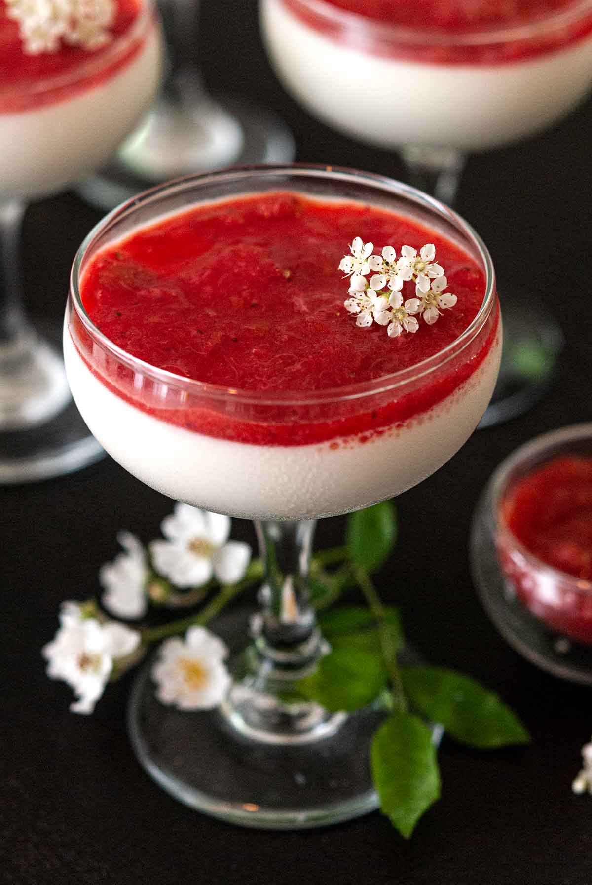 Panna cotta with strawberry-rhubarb compote in a glass, garnished with small flowers, in front of 2 others.