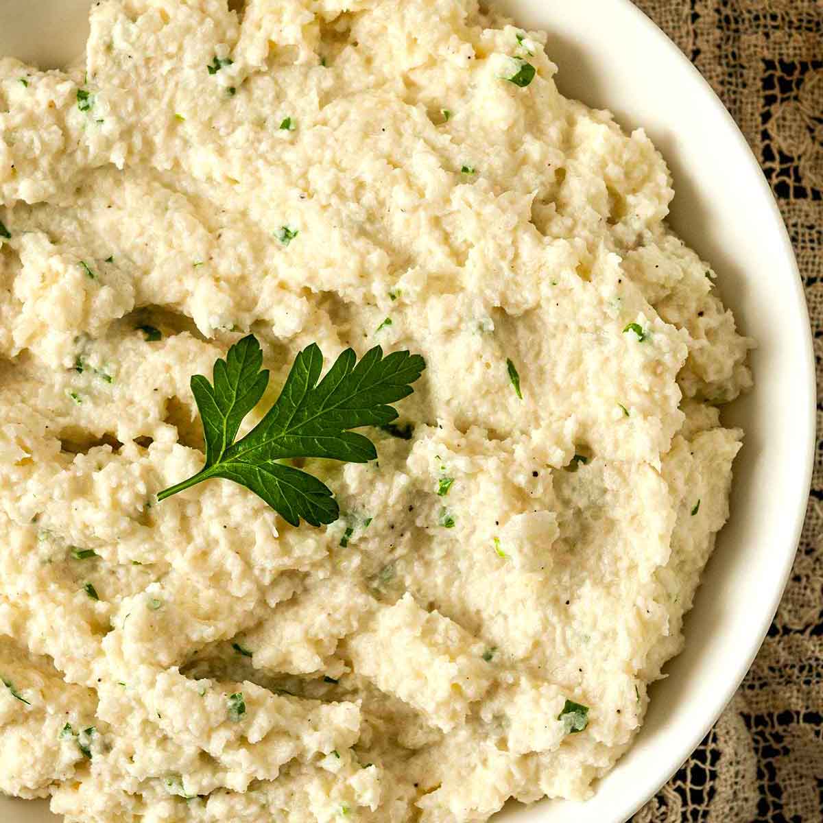 A bowl of garlic-parmesan mashed cauliflower, garnished with a parsley leaf on a lace table cloth.